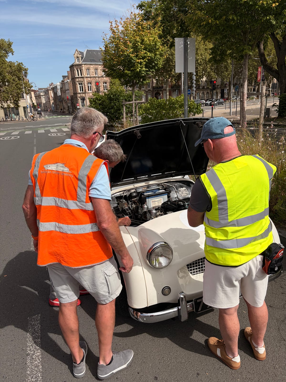 Three men in hi-vis vests peering into TS2's engine bay from the front, the bonnet up and a stone town house across the junction behind
