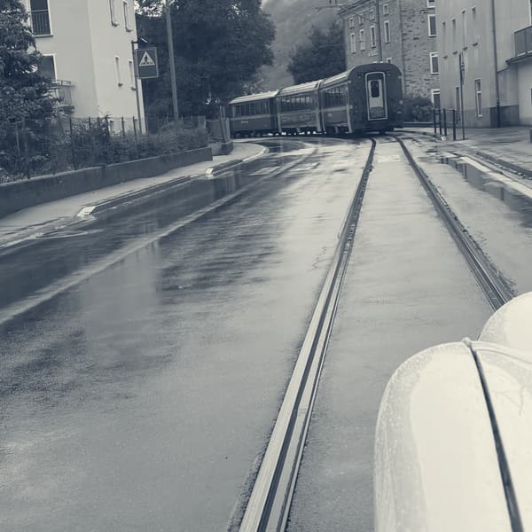 Railway lines running down the middle of a wet village street, the tail of a red train disappearing between the houses ahead