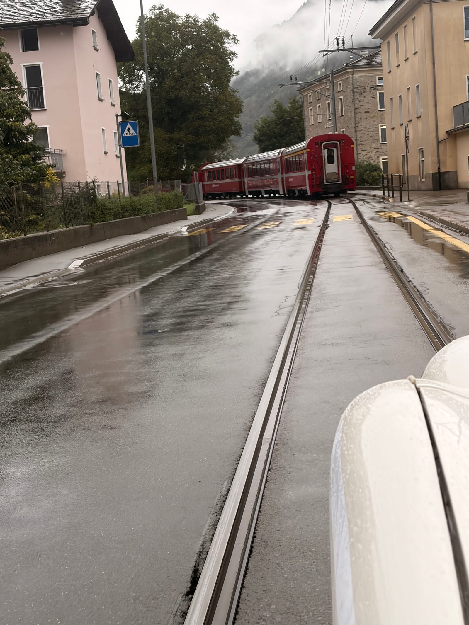 Railway lines running down the middle of a wet village street, the tail of a red train disappearing between the houses ahead
