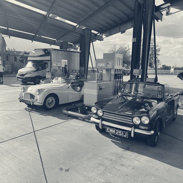 TS2 under a service station canopy beside a maroon Triumph convertible, both nose in at the pumps