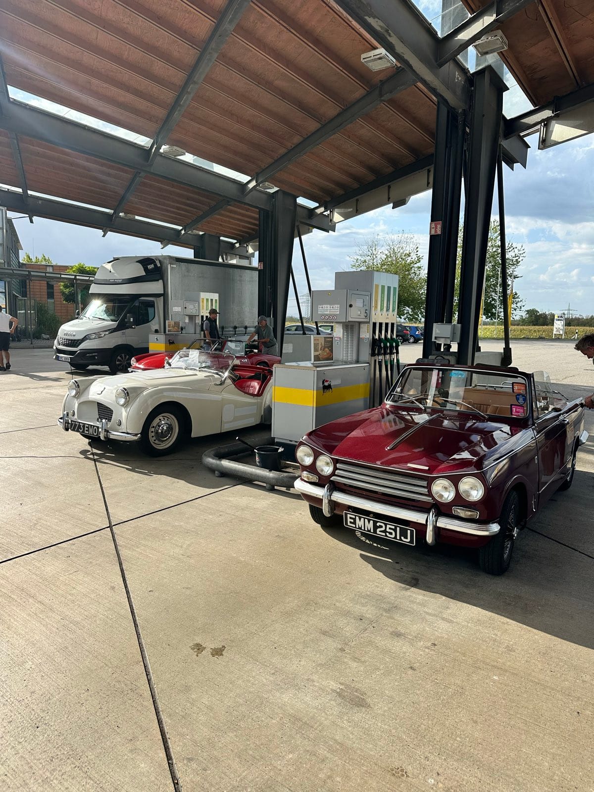 TS2 under a service station canopy beside a maroon Triumph convertible, both nose in at the pumps