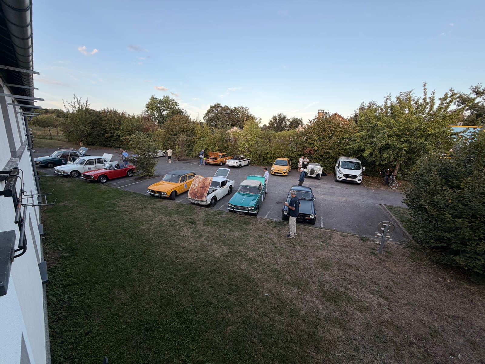 Triumphs parked in rows on a hotel car park at first light, seen from an upstairs window, people standing about between them