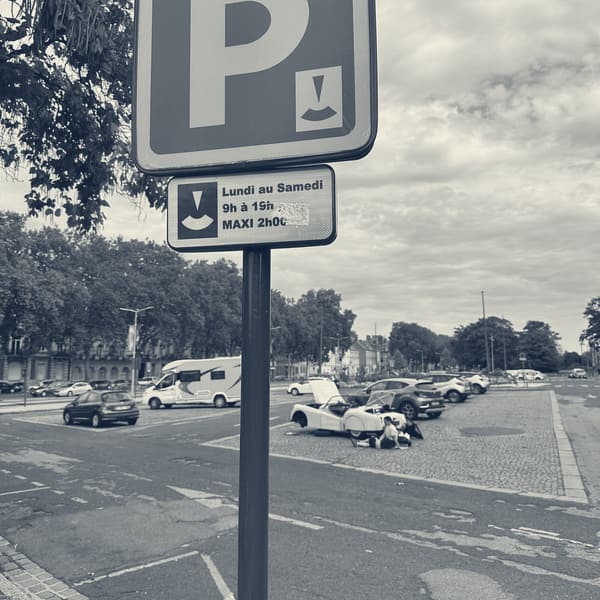 A French blue parking sign with a plate below reading Lundi au Samedi, 9h a 19h, MAXI 2h00, TS2 small in the square beyond with her bonnet and boot open