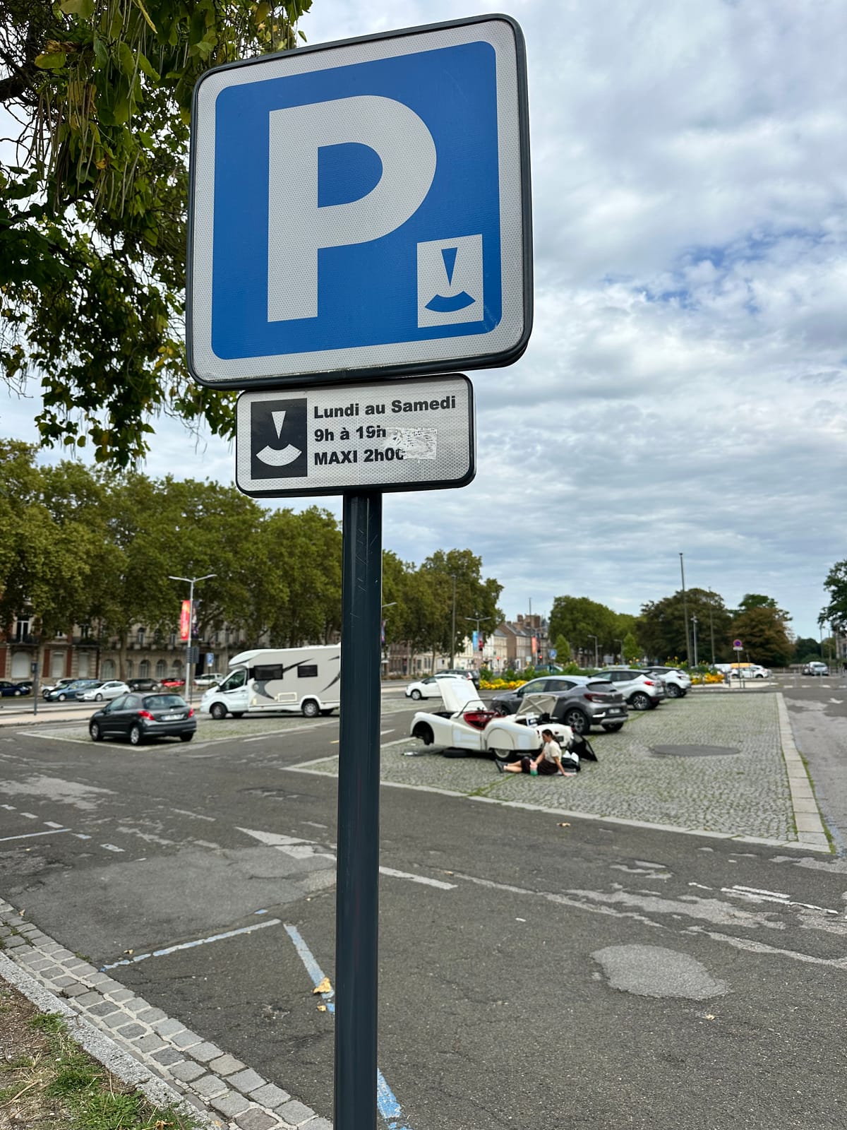 A French blue parking sign with a plate below reading Lundi au Samedi, 9h a 19h, MAXI 2h00, TS2 small in the square beyond with her bonnet and boot open