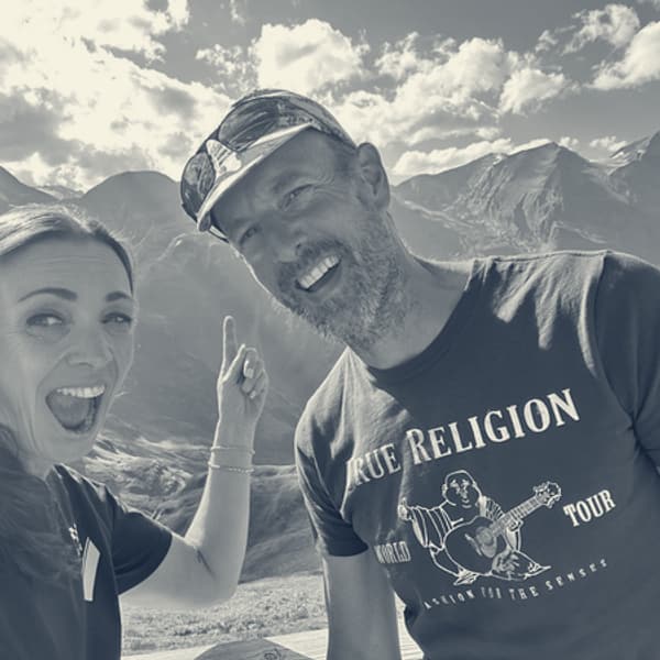 Evie and Andy grinning into the camera at the summit, Evie pointing upward, bare grey peaks behind them