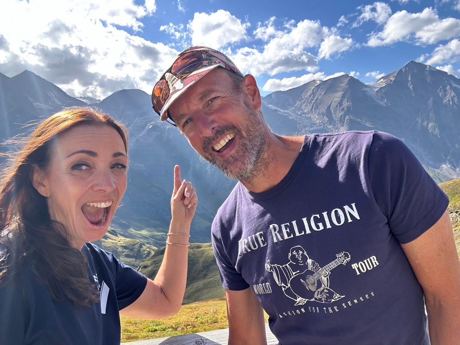 Evie and Andy grinning into the camera at the summit, Evie pointing upward, bare grey peaks behind them
