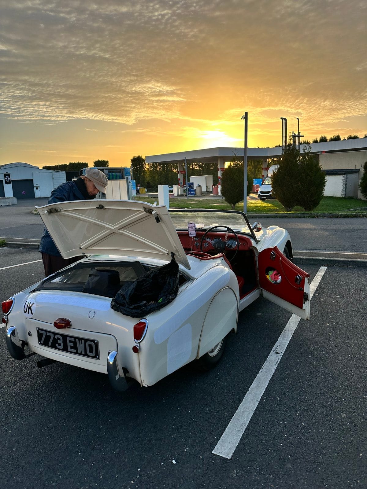 TS2 at Aldi with her boot lid up and Evie leaning into it, the sun coming up orange behind the canopy