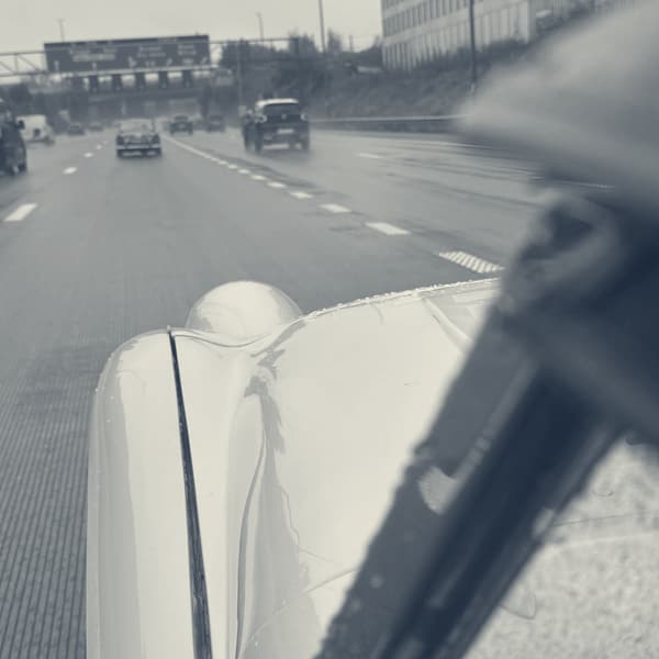 A red TR ahead on a wet grey motorway under blue gantry signs, seen past TS2's rain-beaded white wing and wiper