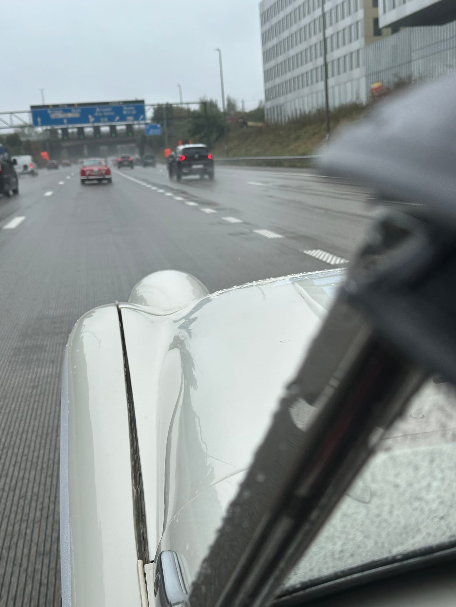 A red TR ahead on a wet grey motorway under blue gantry signs, seen past TS2's rain-beaded white wing and wiper
