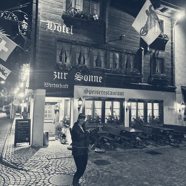 The timbered front of Hotel zur Sonne in Andermatt at night, lamps lit, Evie crossing the cobbles towards the door