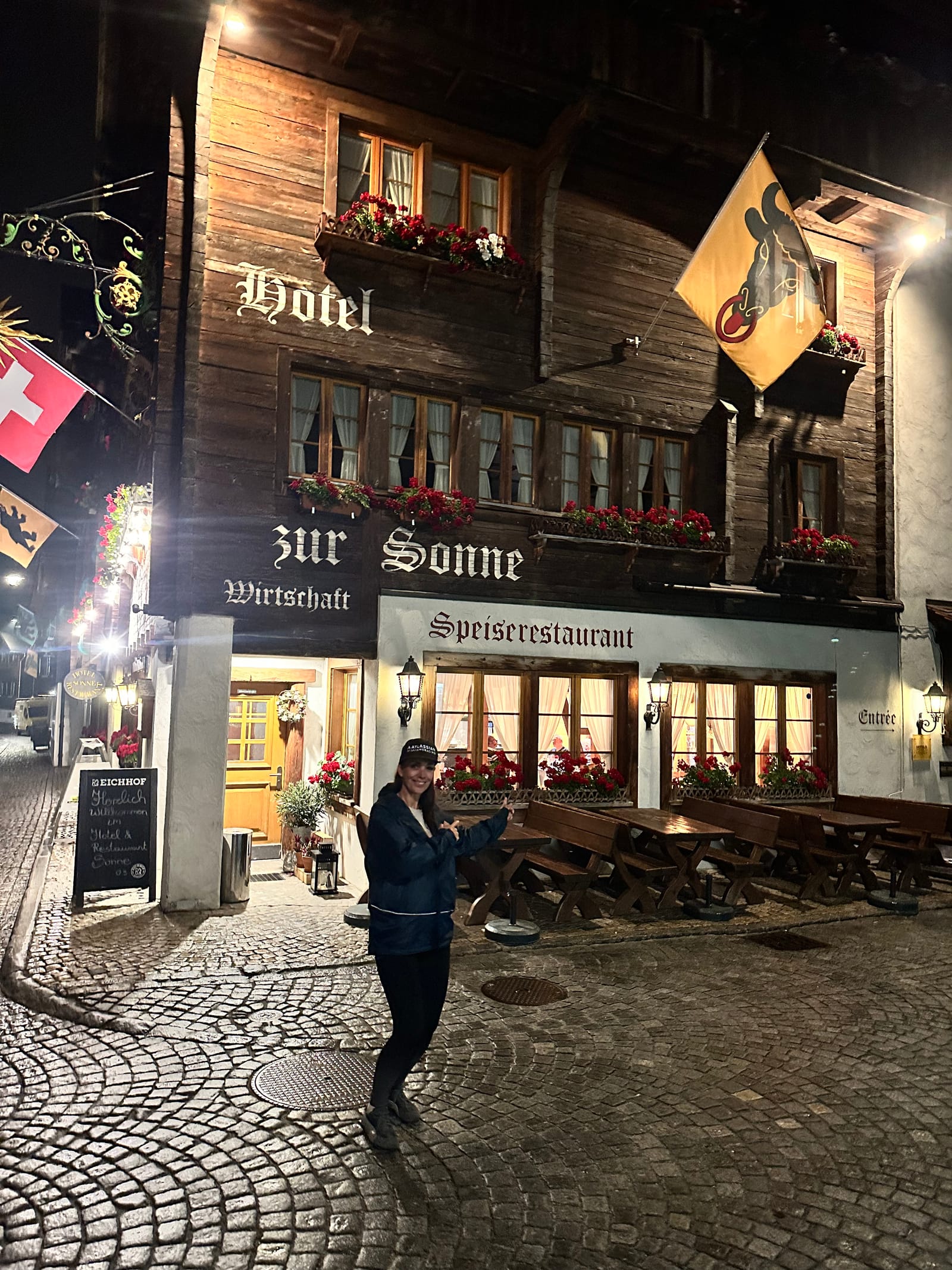 The timbered front of Hotel zur Sonne in Andermatt at night, lamps lit, Evie crossing the cobbles towards the door