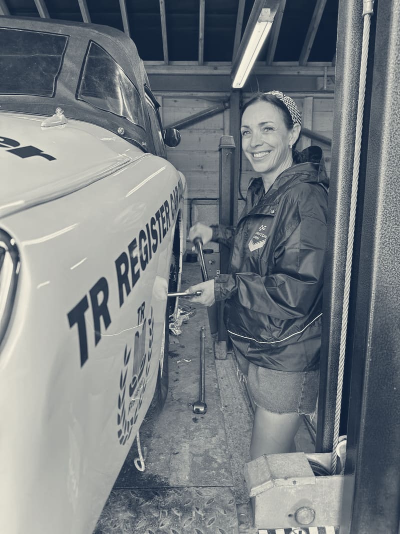 Evie beside TS2 in a garage, the car up on a four post lift with the roof on, torquing the front wheel