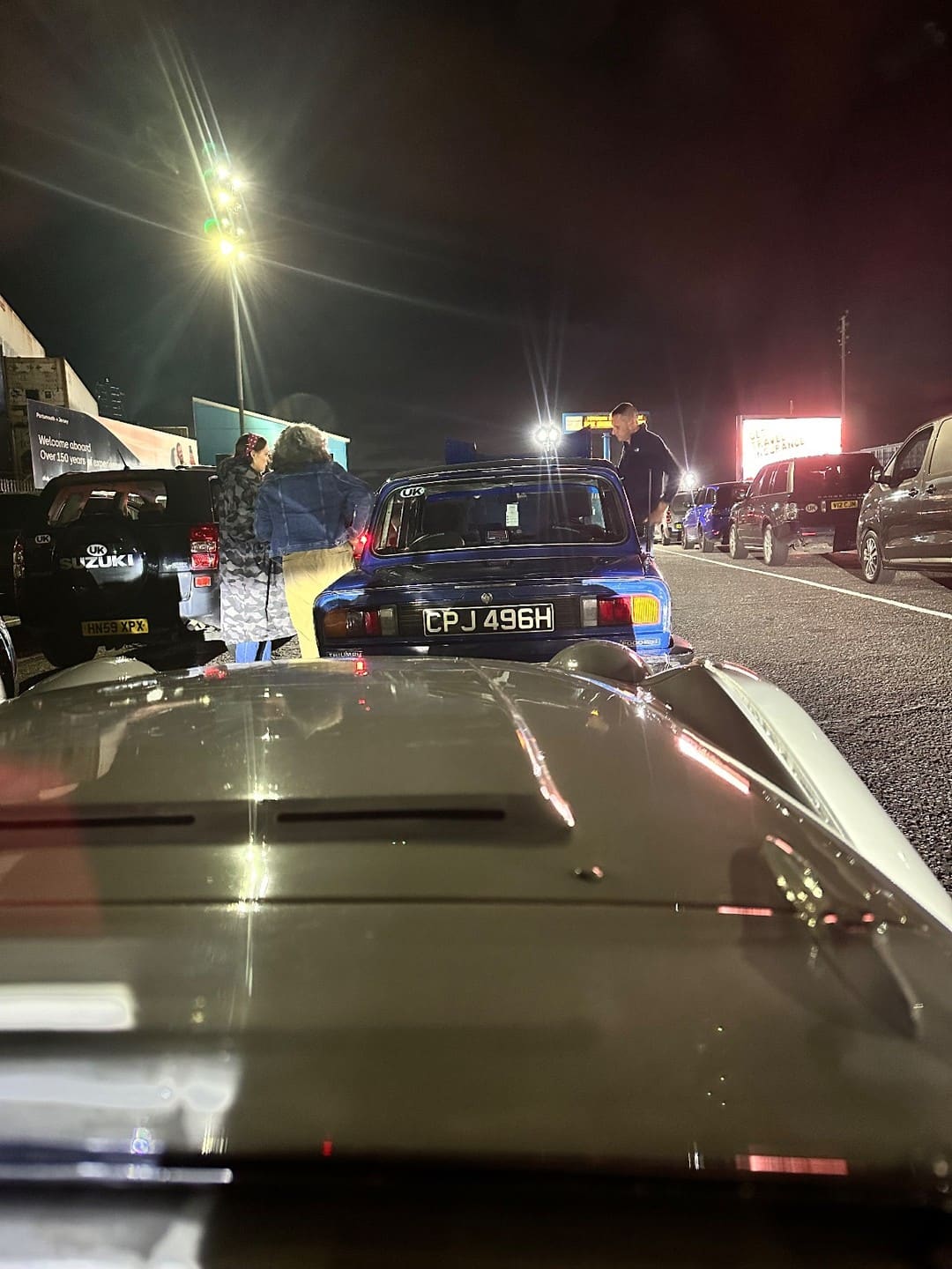 TS2's bonnet in the foreground of the night-time ferry queue at Portsmouth, a blue Triumph 2000 ahead of her under the floodlights